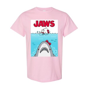 May include: Light pink t-shirt featuring a graphic of a shark with a red bow and a cartoon character in the water. The word "JAWS" is printed in red above the characters. The shirt is a short-sleeved crew neck.