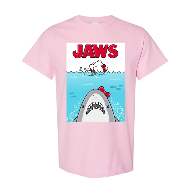 May include: Light pink t-shirt featuring a graphic of a shark with a red bow and a cartoon character in the water. The word "JAWS" is printed in red above the characters. The shirt is a short-sleeved crew neck.