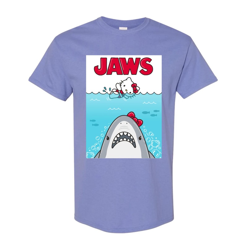 May include: A light purple t-shirt featuring a parody of the movie "Jaws." The graphic shows a cartoon shark with a red bow tie and a cartoon character in the water. The word "JAWS" is printed in red at the top.