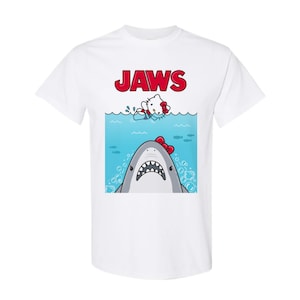 May include: White t-shirt featuring a cartoon graphic of a shark with a red bow tie and a cartoon cat in the water, with the word "JAWS" in red above. The shark has a wide open mouth with sharp teeth.