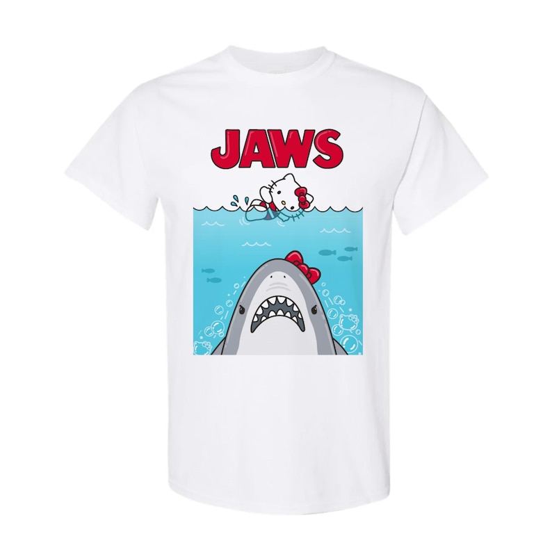May include: White t-shirt featuring a cartoon graphic of a shark with a red bow tie and a cartoon cat in the water, with the word "JAWS" in red above. The shark has a wide open mouth with sharp teeth.