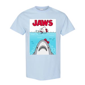 May include: Light blue t-shirt featuring a graphic of a shark with a red bow and a cartoon character in the water. The word "JAWS" is printed in red above the characters. The shirt is made of a soft material.