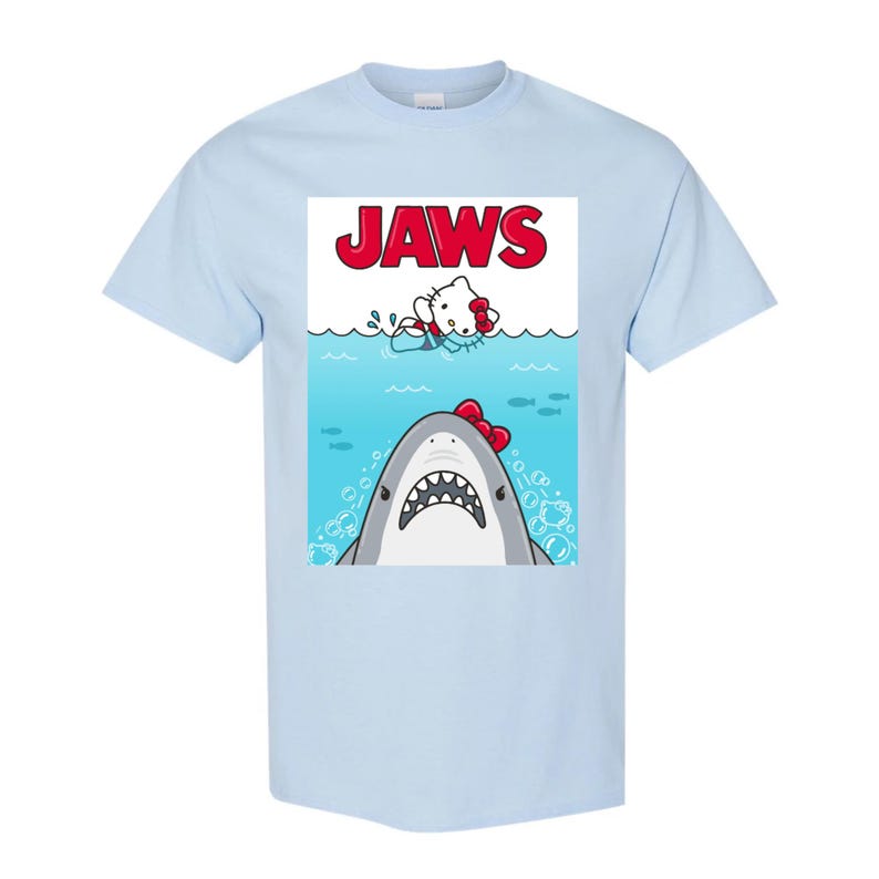 May include: Light blue t-shirt featuring a graphic of a shark with a red bow and a cartoon character in the water. The word "JAWS" is printed in red above the characters. The shirt is made of a soft material.