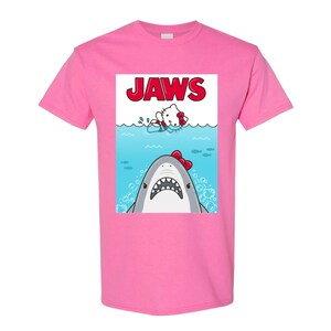 May include: Pink t-shirt featuring a graphic of a shark with a red bow and a cartoon character in the water, with the word "JAWS" in red letters above. The design is a parody of the movie poster.