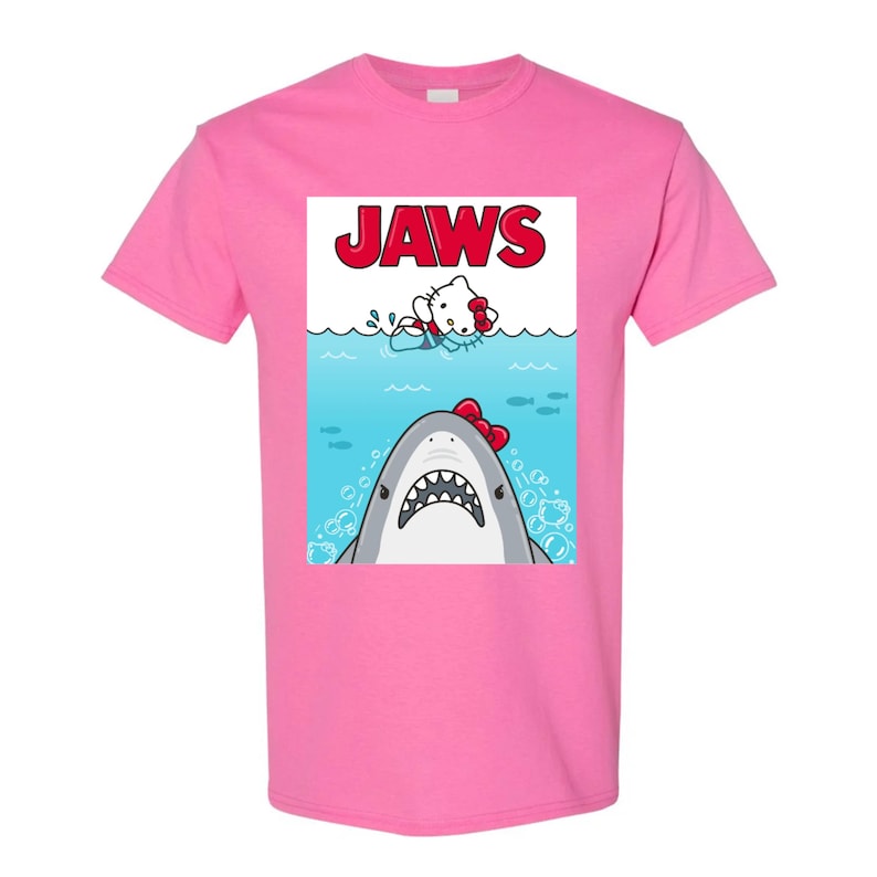 May include: Pink t-shirt featuring a graphic of a shark with a red bow and a cartoon character in the water, with the word "JAWS" in red letters above. The design is a parody of the movie poster.