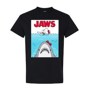 May include: Black t-shirt featuring a parody design. The graphic depicts a shark with a red bow and a cartoon character in the water, with the word "JAWS" in red block letters above. The design is a humorous take on the classic movie poster.