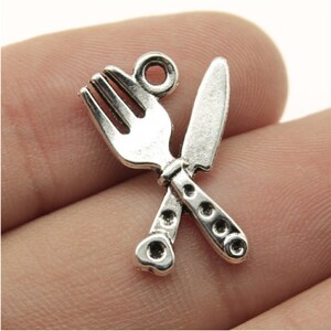 May include: Silver-toned metal charm featuring a crossed fork and knife, perfect for adding a culinary touch to jewelry or crafts.