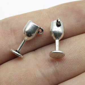 Könnte beinhalten: Zwei silberfarbene Weinglas-Anhänger aus Metall. Jeder Anhänger hat oben eine Schlaufe zum Anbringen an einer Halskette oder einem Armband.