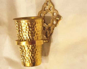 May include: A gold-toned wall mounted toothbrush holder with a decorative design. The holder has a hammered metal finish and a round cup.