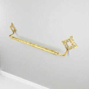 Puede incluir: Una barra de toalla dorada con piezas decorativas en forma de diamante. La barra está montada en una pared gris claro. La barra tiene una superficie texturizada.