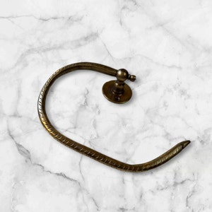 May include: A brass towel ring with a decorative, textured design. The ring is attached to a wall mount with a round base.