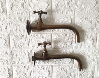 May include: Two antique brass wall-mounted faucets with cross-shaped handles. The faucets have a curved spout and a round base.