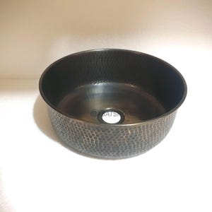 May include: A hammered bronze-colored sink with a round basin and a textured exterior. The sink has a drain hole in the center and the text "IBABATSHOP" is visible. The sink is designed for bathroom or kitchen use.