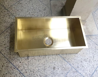 May include: A rectangular, gold-colored kitchen sink with a drain hole. The sink has a polished finish and the text "IBABATSHOP" is visible on the interior. The sink is made of metal and is set against a tiled floor.