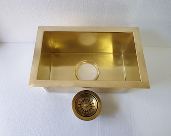 May include: A rectangular kitchen sink with a polished gold-toned finish and a smooth metallic surface. The sink features a central circular drain opening and is accompanied by a matching circular gold-toned drain strainer assembly.