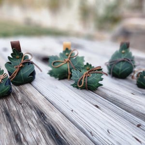 May include: A collection of green leaf-shaped leather pouches with brown leather ties and accents. The pouches vary in size and are arranged on a weathered wooden surface. Some have a small brown leather loop.