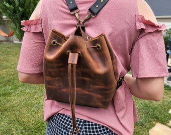 May include: A brown leather bucket bag with a drawstring closure and adjustable straps. The bag is worn by a person and has a distressed look.