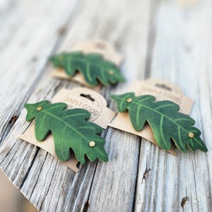 May include: Three green leather leaf-shaped items with brass rivets are displayed on a weathered wooden surface. Each leaf is attached to a small, brown paper tag with the words "SIMPLE GOOD" printed on it. The leaves have detailed vein patterns.