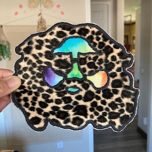 May include: A leopard print patch with a colorful tie-dye design in the center. The patch has a black border and is held by a hand. The tie-dye colors include blue, green, purple, and orange.