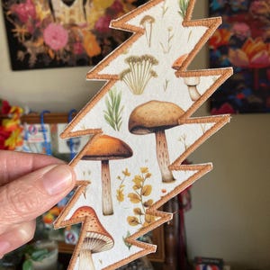 May include: A white fabric patch in the shape of a lightning bolt, featuring a detailed illustration of various mushrooms and foliage. The patch has a brown border and showcases a botanical theme.