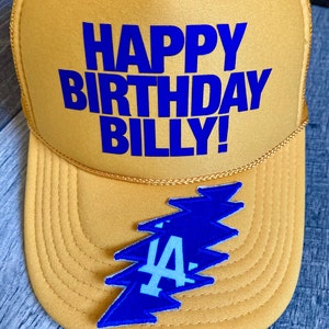 May include: A yellow trucker hat with a blue "Happy Birthday Billy!" inscription and a blue lightning bolt patch with a white "LA" inside.