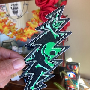 May include: Green and black embroidered patch with a skull and crossbones design. The patch is shaped like a lightning bolt and has a zig-zag border.