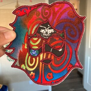 May include: A colorful, embroidered patch with a unique design. The patch features a face-like image with a skull, swirls, and floral elements in red, blue, purple, and green. The patch is outlined in a dark red color.