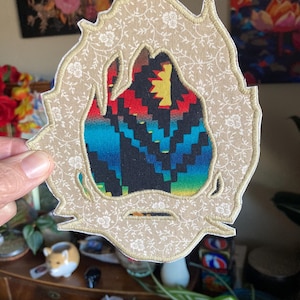 May include: An embroidered patch with a flame-like shape. The patch features a beige floral patterned border with a colorful geometric design in the center, including red, blue, and yellow. The patch is held by a hand.