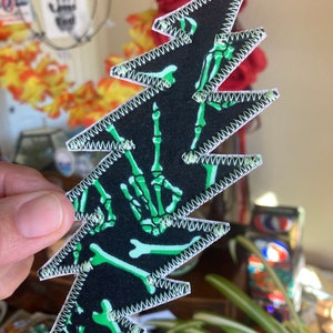 May include: Green skeleton hand print patch with white stitching on a black background. The patch is shaped like a lightning bolt.