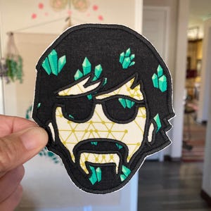 May include: Embroidered patch featuring a stylized face with black hair and sunglasses. The face has a cream-colored background with a geometric pattern. Green crystal-like shapes are scattered throughout the hair and beard. The patch has a black border.