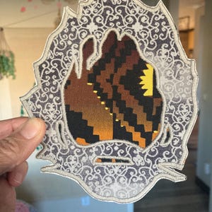 May include: An embroidered patch with a decorative border. The patch features a central design with brown, black, and yellow geometric shapes. The border is a detailed, swirling pattern in shades of gray and white.