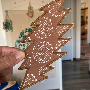 May include: Brown lightning bolt patch with white floral pattern and brown border. The patch is held up in front of a blurred background. The lightning bolt design is a popular symbol.