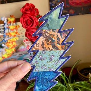 May include: A multi-colored lightning bolt patch with a blue embroidered border. The patch is made of fabric with various patterns, including floral and batik designs. The patch is held up in front of a background with flowers and plants.