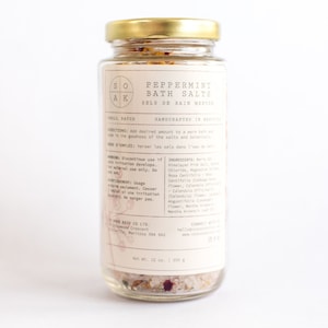 Puede incluir: Un frasco de vidrio con tapa dorada lleno de sales de baño. La etiqueta dice "Sales de baño de menta" y "Hecho a mano en Manitoba". La lista de ingredientes incluye sal rosa del Himalaya, cloruro de magnesio, rosa centifolia, caléndula officinalis y hoja de menta arvensis.