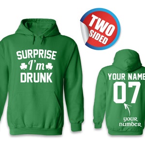 May include: Green hooded sweatshirt with the text "Surprise I'm Drunk" and shamrock symbols on the front. The back of the sweatshirt has the text "Your Name" and the number "07" in white.