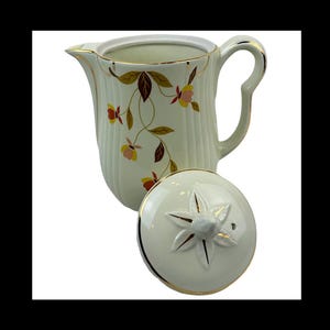 May include: A vintage cream-colored ceramic creamer and lid. The creamer features a ribbed design and is adorned with a floral pattern in shades of yellow, brown, and pink. Gold trim accents the rim, handle, and lid.