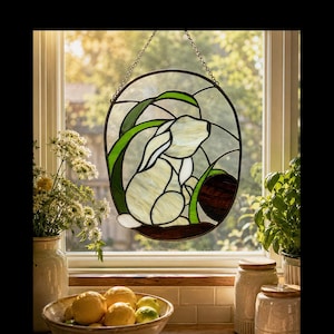 May include: An oval stained glass suncatcher featuring a white rabbit sitting among green leaves and a dark brown circular element. The piece is framed in black metal and suspended by a silver chain, displaying a translucent, multi-colored design.