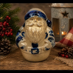 May include: A ceramic Santa Claus figurine featuring a cream-colored body with a speckled texture and deep blue painted details on the hat, belt, and coat accents. The figure has a prominent beard, mustache, and a rounded, stout physical form.