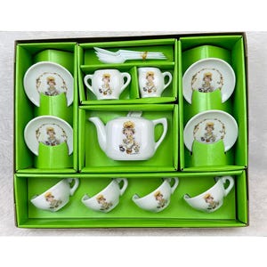 May include: A white porcelain tea set featuring a central teapot, a sugar bowl, a creamer, four teacups, four saucers, and four small spoons. Each piece is decorated with a graphic illustration of a child with brown hair and a yellow hat.