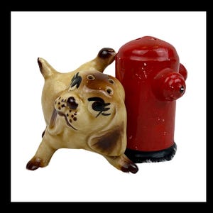 May include: A ceramic salt and pepper shaker set featuring a tan dog with brown ears and spots standing next to a bright red fire hydrant. The dog has painted black eyes and eyelashes, while the red fire hydrant has a black base.