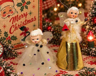 May include: Two vintage-style angel ornaments with painted faces and blonde hair. One angel wears a white tulle skirt with colorful sequins and holds a red flower. The other wears a fuzzy white top, a gold cone skirt, and holds a red bell.