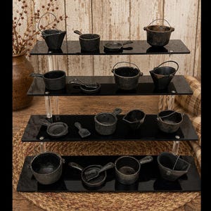 May include: A collection of miniature black cast iron cookware pieces, including small pots, pans, buckets, and ladles, arranged on a four-tiered black acrylic display stand. Each piece features a dark, textured finish and metal handles.