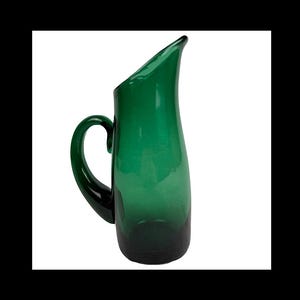 May include: A green glass pitcher with a curved handle and a slanted spout. The pitcher has a smooth, glossy finish and a dark green base that fades to a lighter green towards the top. This decorative pitcher is ideal for serving beverages.
