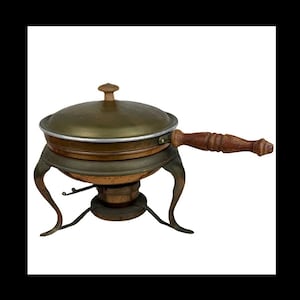 May include: An antique copper and brass chafing dish with a wooden handle. The dish has a lid with a small knob and sits on a decorative stand with curved legs. The chafing dish is designed to keep food warm.