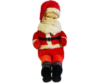 May include: A vintage plush Santa Claus doll wearing a red suit with white trim, a black belt, and a white beard. He has a painted face with black eyes and a red nose.