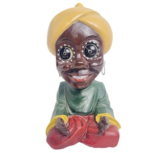 May include: A figurine of a person with brown skin, wearing a yellow turban, green and red clothing, and large eyes with black pupils.