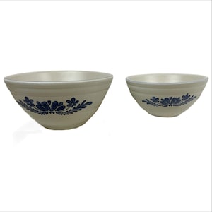 May include: Two off-white ceramic bowls with a blue floral design. The bowls have a rounded shape and a blue trim around the rim. The larger bowl is on the left, and the smaller bowl is on the right. The floral design is repeated on both bowls.