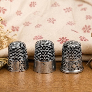 May include: Three antique silver thimbles with intricate designs, arranged on a wooden surface. The thimbles have a mesh-like top and decorative bands. A spool of thread and a pair of scissors are also visible, suggesting a sewing theme.