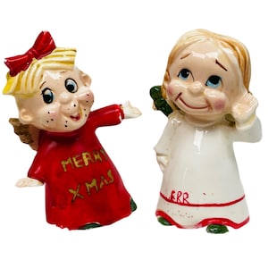 May include: Two ceramic Christmas angel figurines. One angel is wearing a red dress with gold lettering that says "Merry Xmas". The other angel is wearing a white dress with red trim and gold lettering that says "Merry".