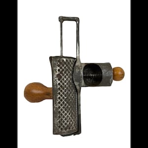 May include: A vintage metal nutmeg grater with a wooden handle and a rectangular grating surface. The grater has a handle on top for easy use. The wooden handle is a light brown color.